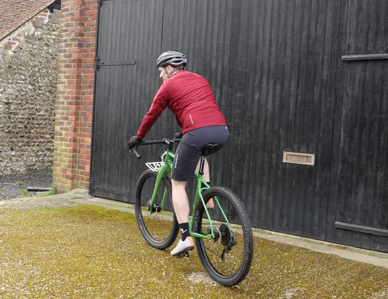 Rear view of the GORE R5 WINDSTOPPER shirt worn on a green bike, rider heading across a yard past black barn doors.