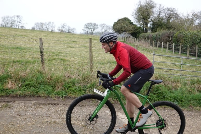 Rider in the GORE R5 WINDSTOPPER shirt pedals a green bike along a gravel track beside a grass field.