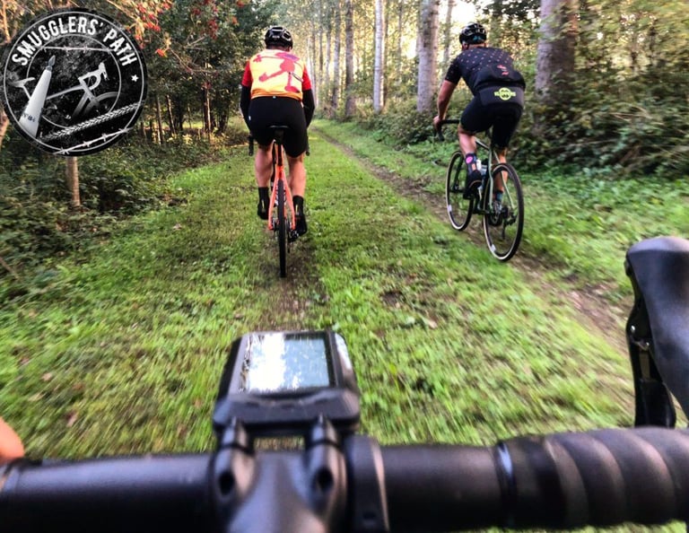Two riders follow a grassy woodland track on the Smugglers Path gravel route in Belgium, seen from the handlebars.