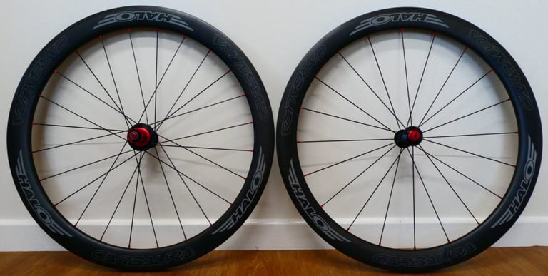 Halo Carbaura RC carbon wheelset with red hubs and black spokes, standing against a white wall on a wooden floor.