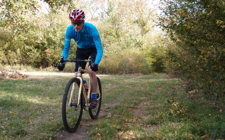 Rider in a blue jersey and red helmet rides a cream gravel bike on Halo Vapour GXC wheels along a grassy autumn trail.