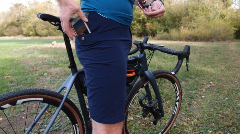 Rider slides a phone into a popper-fastened rear pocket of navy Café du Cycliste Renée shorts beside a gravel bike.