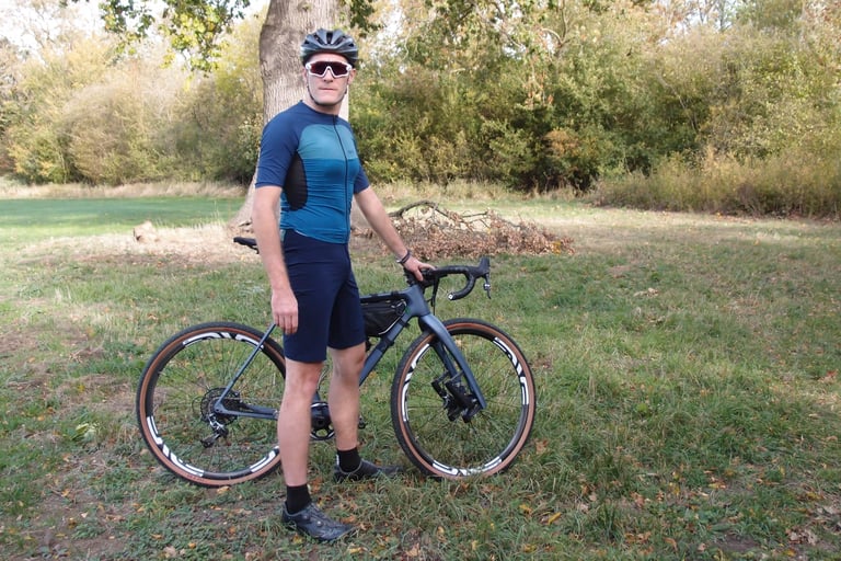 Rider in Café du Cycliste Renée Gravel Shorts, helmet and sunglasses stands holding a gravel bike on grass.