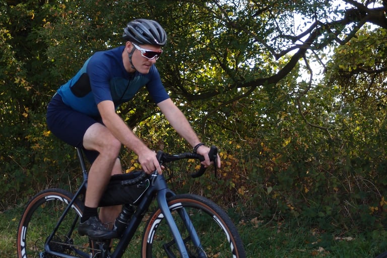Side view of a rider pedalling a gravel bike in the Café du Cycliste Renée shorts past autumn hedgerow.