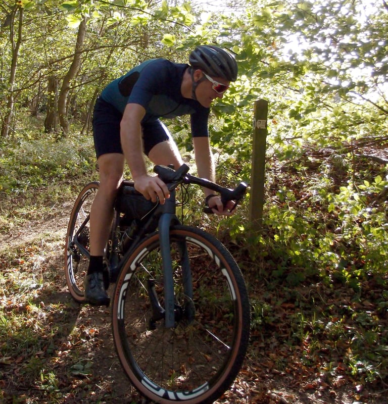 Rider in Café du Cycliste Renée Gravel Shorts and a teal jersey rides a gravel bike along a wooded trail.