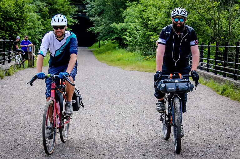 Two bikepackers ride a wide gravel trail towards the camera, one with a bar bag and the other a rear rack bag.
