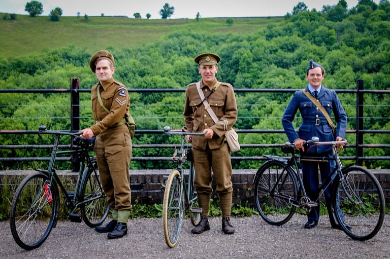 Three men in wartime military uniforms stand with vintage roadster bicycles before a wooded valley.