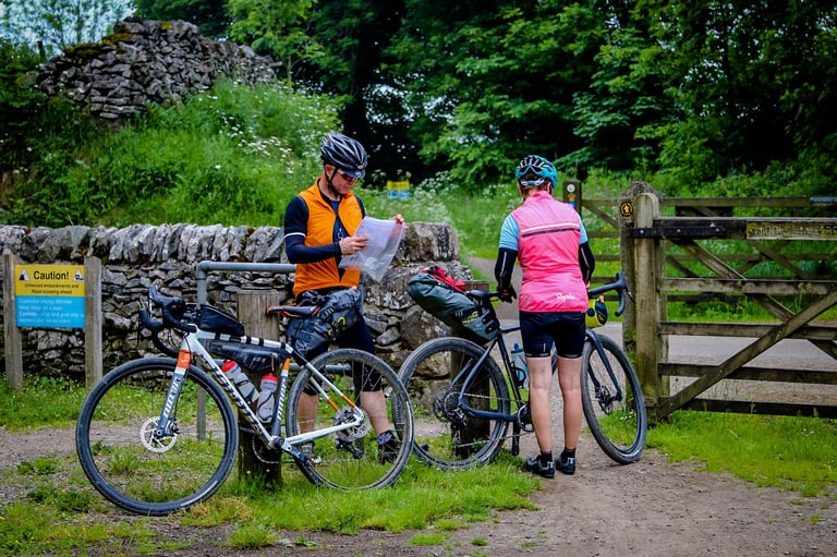 Two loaded gravel bikers stop at a gate, one checking a map, beside a sign warning cyclists to give way.