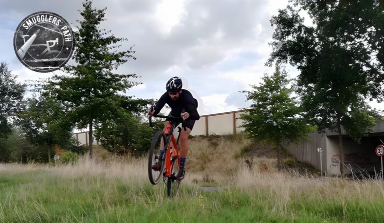 Rider pulls a wheelie on an orange gravel bike on grass beside a road underpass during a Smugglers Path ride.