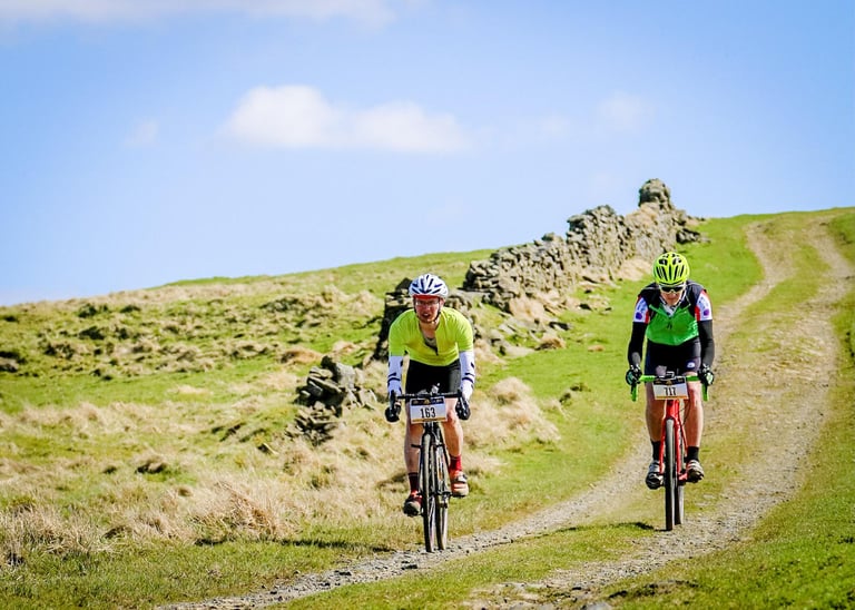 Two gravel riders with race numbers climb a grassy double-track beside a drystone wall under blue sky.