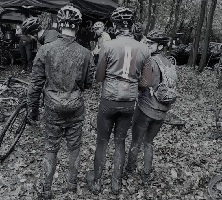 Three mud-spattered riders in winter kit stand among fallen leaves beside a Rapha tent after a wet off-road ride.
