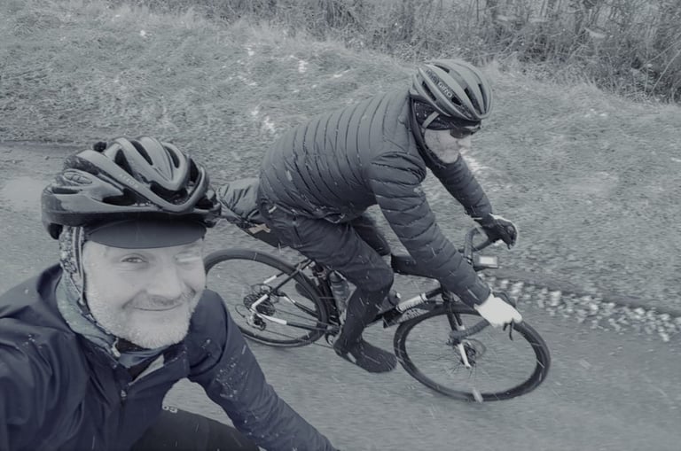 Smiling rider takes a selfie as a second rider in a down jacket cycles alongside on a wet winter lane.