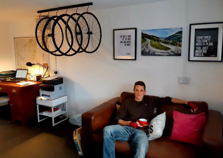 Ben Sharp sits on a leather sofa with a red mug beneath a row of wheels hung from a rail in his workshop.