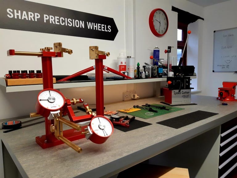 Red wheel truing stand with dial gauges and a spoke cutter on the bench at Sharp Precision Wheels.