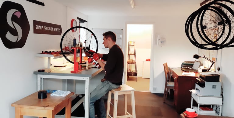 Ben Sharp truing a wheel on a stand in the Sharp Precision Wheels workshop, with the brand logo on the wall.