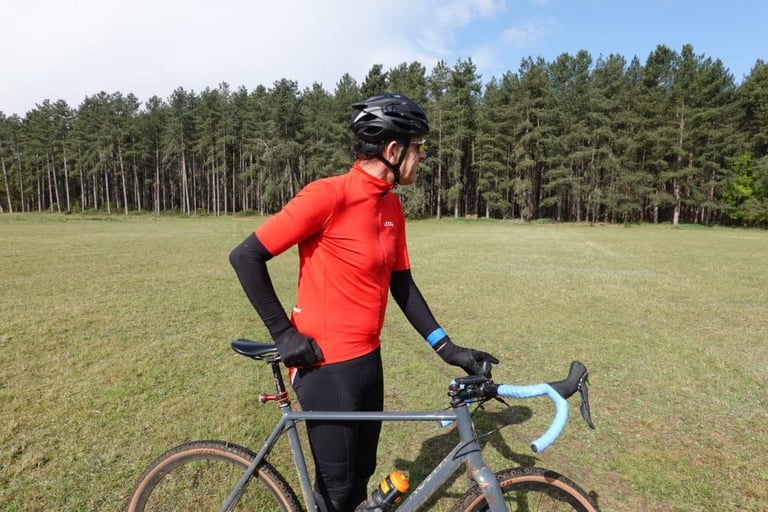 Rider stands with a gravel bike in a field, showing the Le Col Therma Jersey in profile with arm warmers.