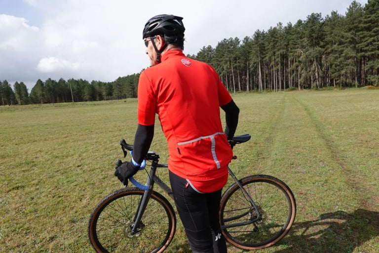 Rear of the red Le Col Therma Jersey with reflective trim, rider standing beside a gravel bike in a field.