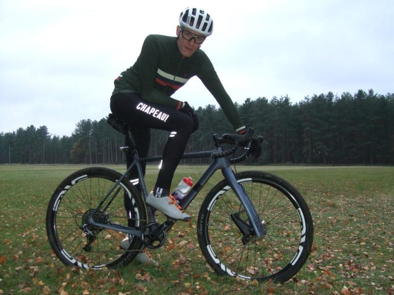 Rider in the Chapeau Club Thermal Bibtight and a green jersey stands astride a gravel bike on a leaf-strewn field.