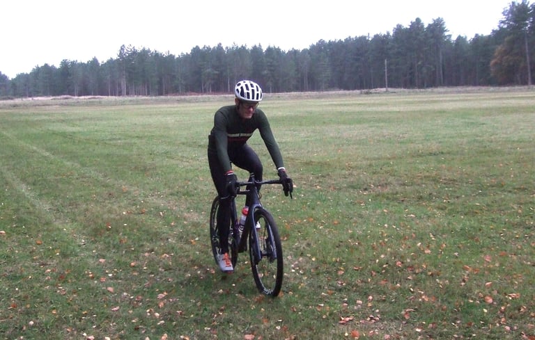 Rider tests the Chapeau Club Thermal Jersey on a gravel bike across an open grassy field edged with pines.