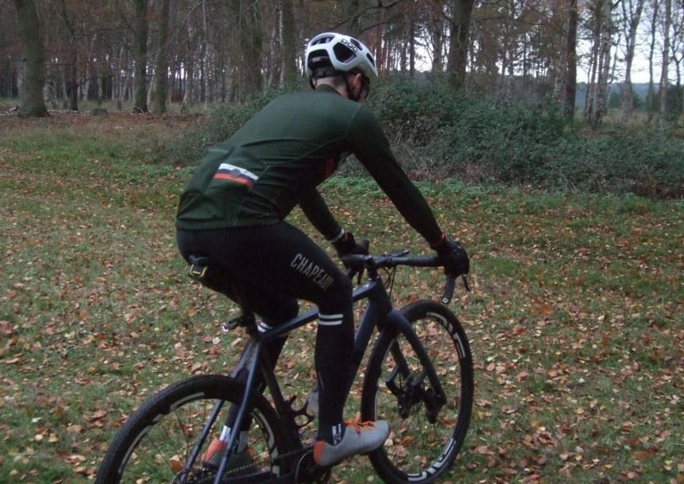 Rider in Chapeau thermal bibtights and a green long-sleeve jersey rides a gravel bike across a leaf-covered clearing.
