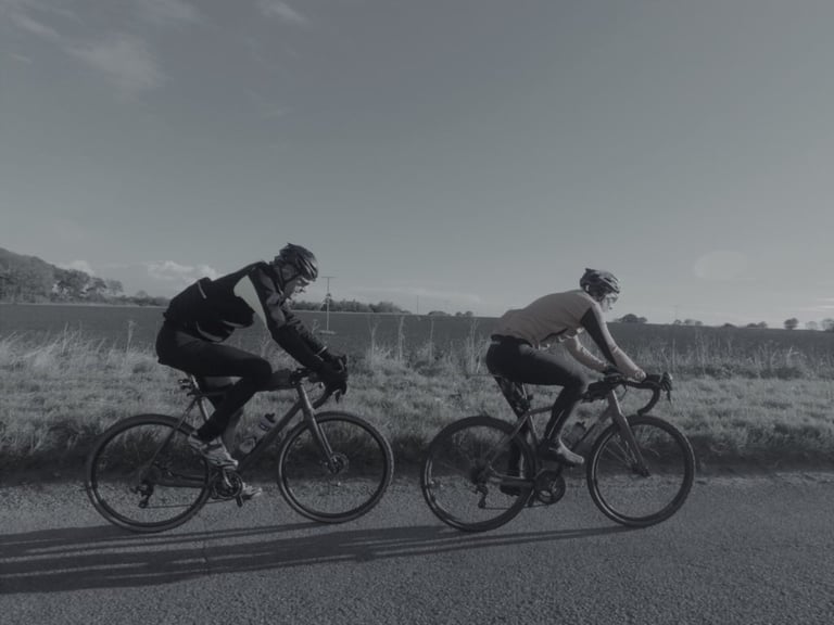 Two riders in winter kit on drop-bar bikes pedal along a quiet country road under a clear sky.