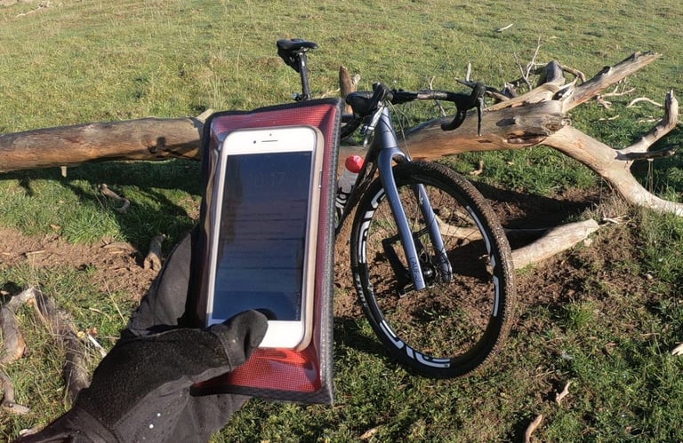 Gloved hand holds a phone in the Fickaskåp wallet beside a gravel bike on grass, screen visible through the window.