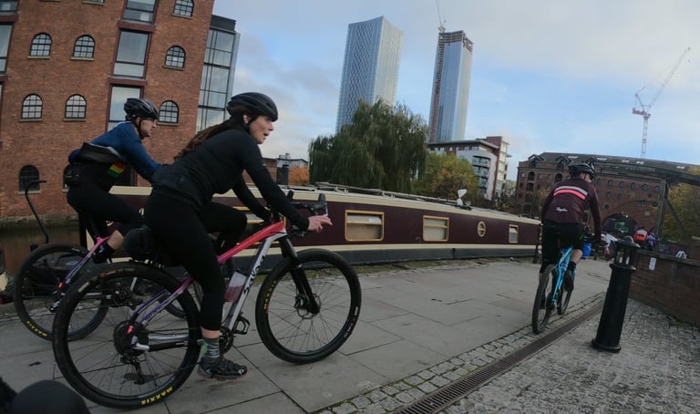 Riders cycle along a Manchester canal towpath past a narrowboat and city towers on the Rapha Prestige.