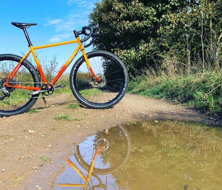 All-City Gorilla Monsoon on a sunlit dirt track, its front wheel and frame reflected in a puddle.