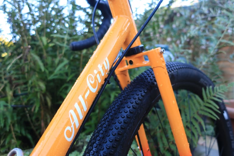 Close-up of the All-City logo on the Gorilla Monsoon's orange down tube, with the steel fork and a knobbly tyre.