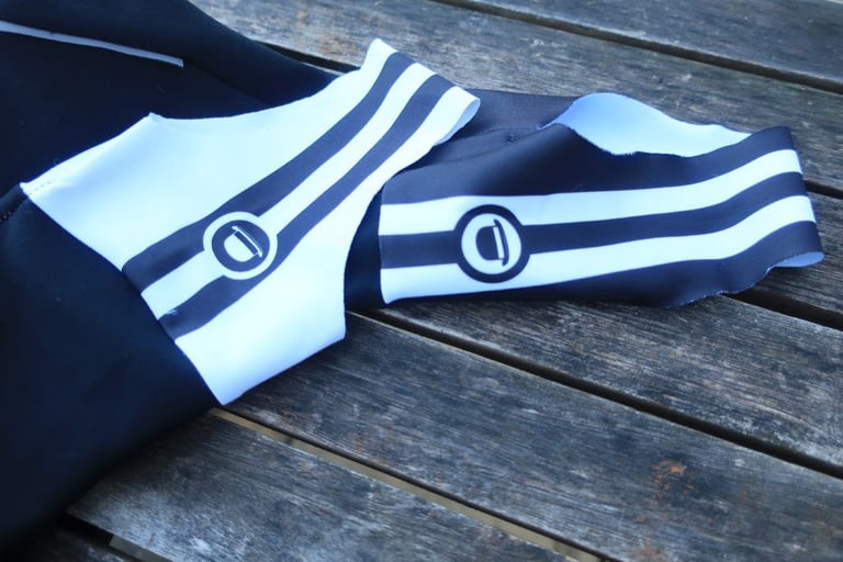 Ankle stirrups of the Chapeau bibtights, white with black stripes and a round logo, laid on a wooden table.
