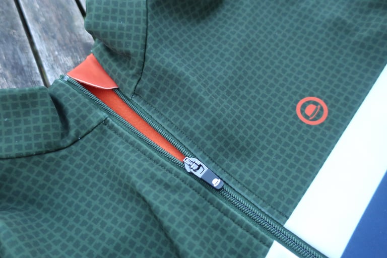 High collar, zip garage and orange lining of the Chapeau Club Thermal Jersey, with a round hat logo.