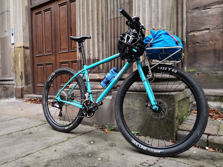 Surly Bridge Club with the 24-pack front rack and a blue dry bag, leaning on a stone doorway with a helmet on the bars.