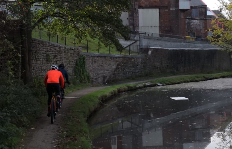 Two riders on a canal towpath past old brick warehouses, one in the orange POC AVIP Ceramic Thermal Jersey.
