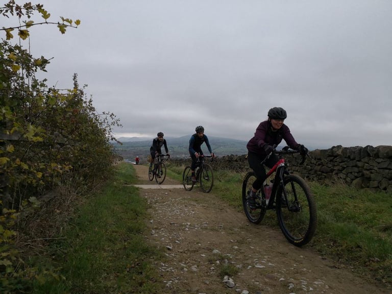 Three riders on a stony farm track beside a drystone wall, with hills behind, on the Rapha Prestige Manchester.