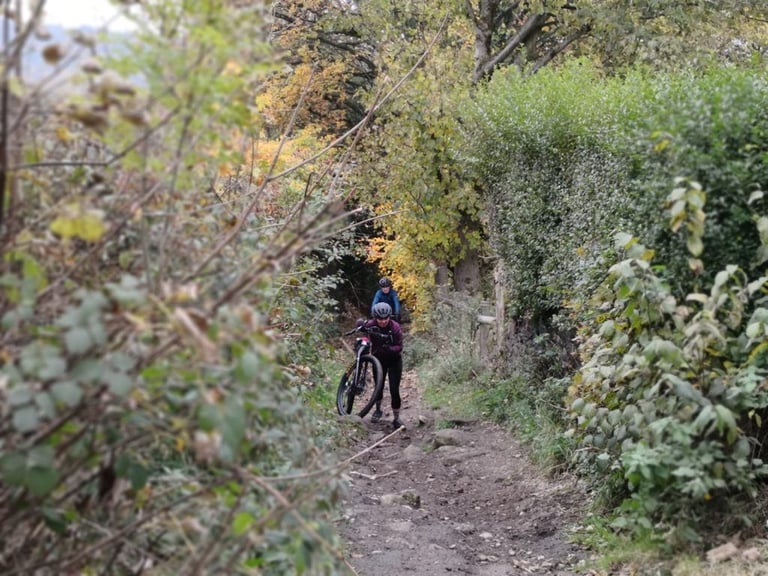 Rider pushes a bike up a narrow, rocky path between autumn hedges on the Rapha Prestige Manchester route.