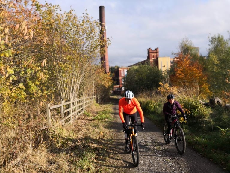Two riders on a gravel path pass a mill chimney on the Rapha Prestige Manchester route towards Stockport.
