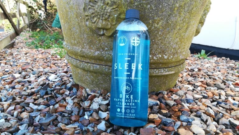 Bottle of Sleek bike cleaner leaning on a moss-covered stone pot among pebbles, shot outdoors in daylight.