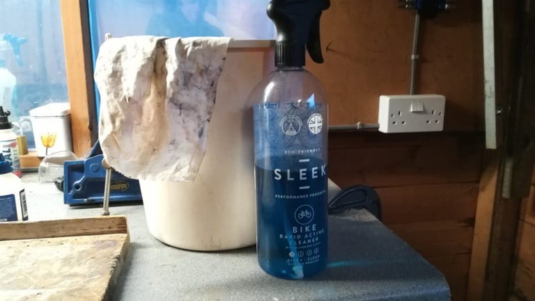 Sleek bike cleaner spray bottle on a workshop bench beside a bucket with a dirty rag draped over it.