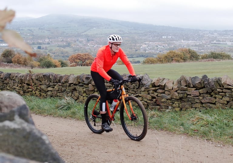 Rider in an orange jersey on the All-City Gorilla Monsoon rides a gravel lane beside a drystone wall, hills behind.