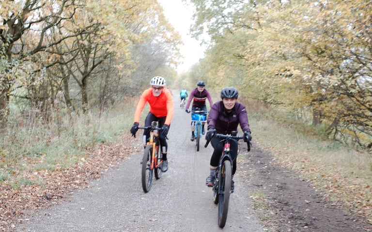 Three riders on gravel bikes roll along a leaf-strewn track between autumn trees on the Rapha Prestige Manchester.