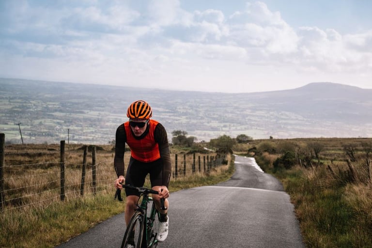 Rider in an orange Victory Chimp Ogham gilet climbs a narrow road above a hazy valley on a green bike.
