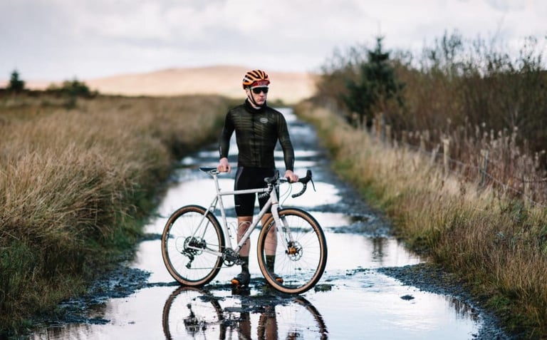 Rider in dark Victory Chimp Ogham kit stands astride a white Donard bike on a puddled moorland track.