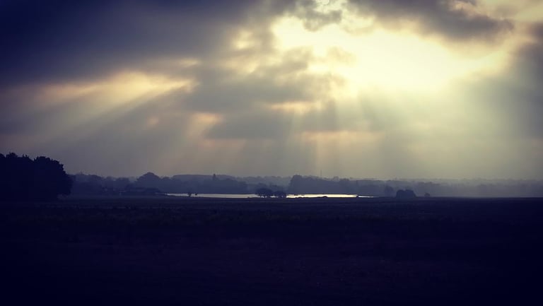 Low winter sun breaks through heavy cloud in rays over dark fields and a distant lake.