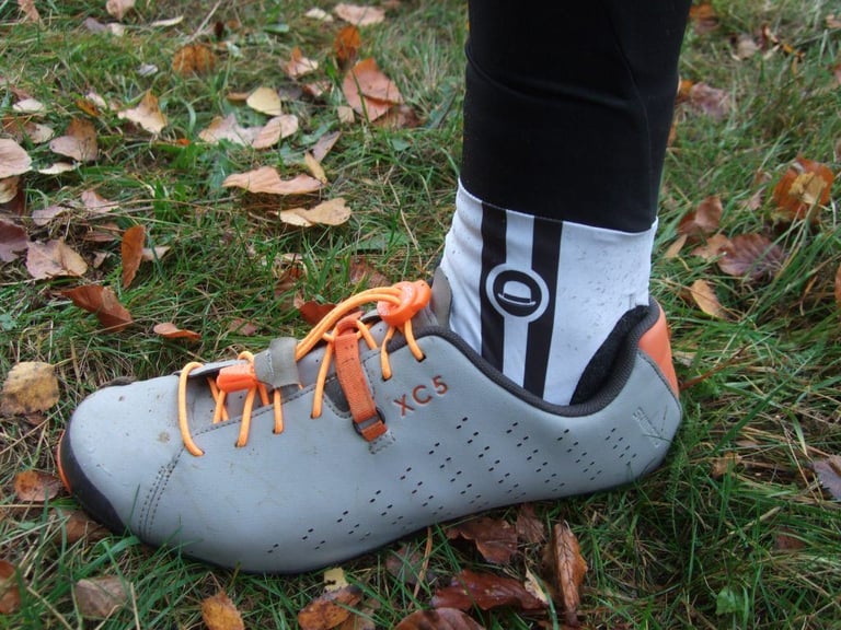 Dirty white Chapeau ankle stirrup over a grey XC5 shoe with orange elastic laces, on a leafy lawn.
