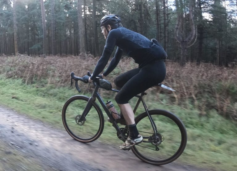 Rider in black kit on a gravel bike along a muddy track in Thetford Forest while testing the Alpkit Comet gloves.