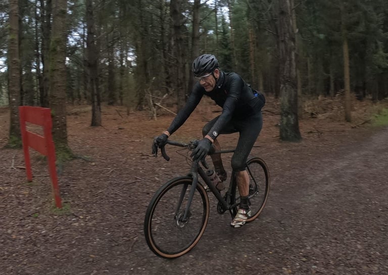 Front view of a rider in mud-spattered Alpkit Rhythm 3/4 Bibtights riding a gravel bike along a forest fire road.
