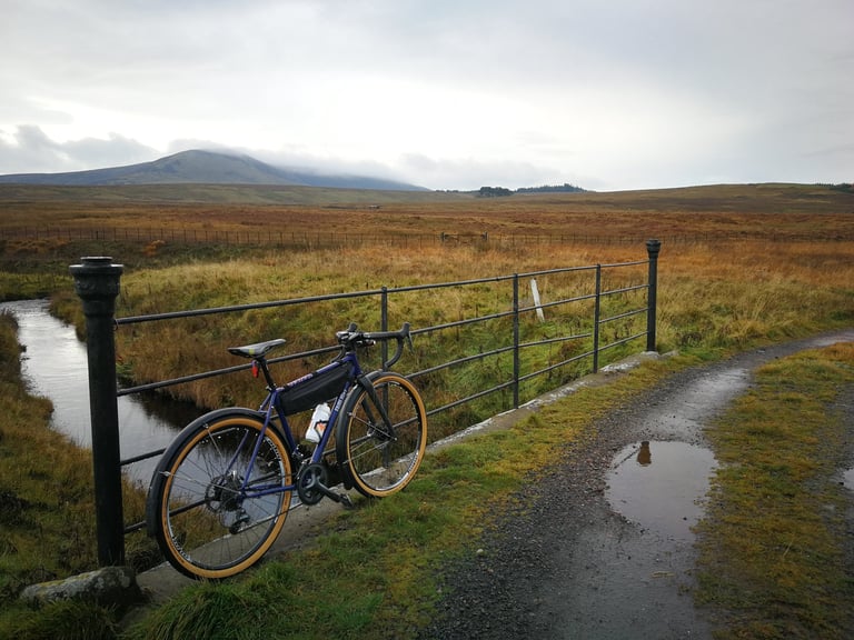 Fairlight Secan leaning on an iron fence beside a moorland stream, with cloud-topped hills behind.