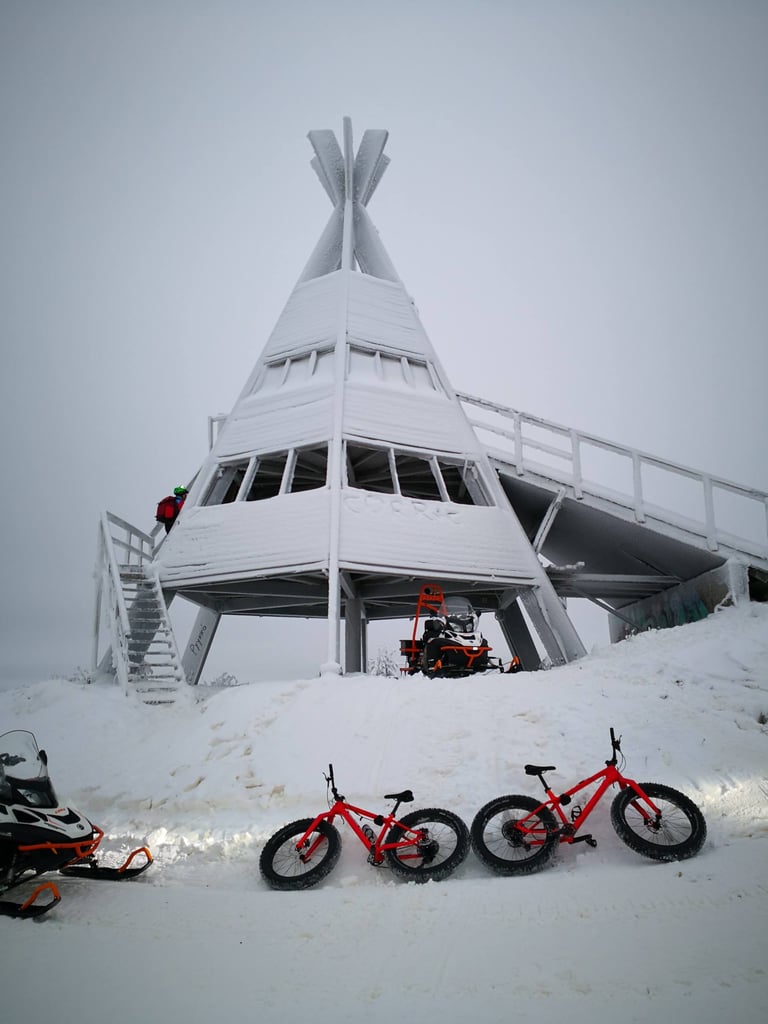 Two red fatbikes in the snow below a white lookout tower, with snowmobiles nearby, on a Roll Outdoors ride in Lapland.