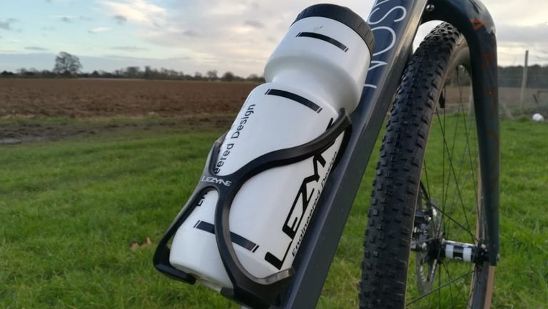 Lezyne Flow Cage holding a white bottle on a gravel bike frame, with a ploughed field behind.