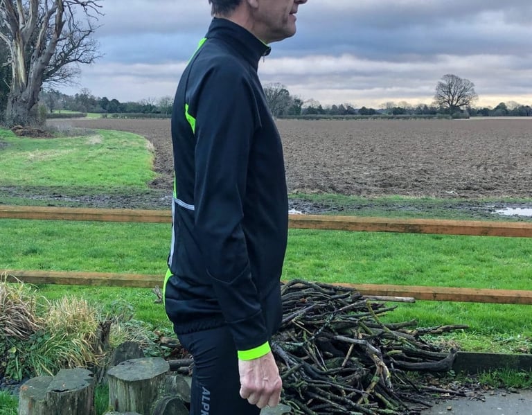 Side view of the black Galibier Mistral jacket, with hi-vis yellow cuff and piping, beside a ploughed field.